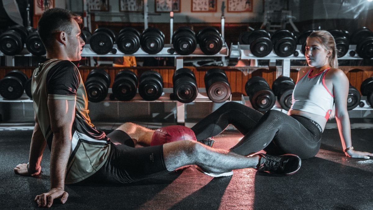 A man and woman resting together on the gym floor, surrounded by dumbbells.